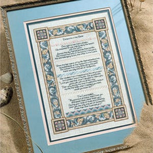 May include: A framed cross-stitch of the poem "Footprints in the Sand." The text is framed by a decorative border with floral and geometric patterns in blue, beige, and pink. The frame is gold-toned, and the artwork is displayed on a light blue background.