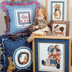 May include: A collection of Easter-themed home decor items. Includes framed cross-stitch pictures of bunnies, decorative pillows with bunny designs, and a stuffed bunny doll. The color palette features blues, pinks, and browns, creating a cozy, country aesthetic.