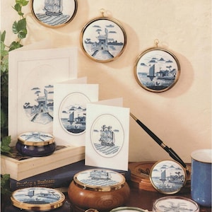 Könnte beinhalten: Eine Sammlung nautischer Kreuzstichkunst und Dekorationsartikel. Enthält gerahmte runde Stücke, kleine runde Boxen, Untersetzer und Grußkarten, alle mit blau-weißen Schiffs- und Küstenmotiven. Die Artikel sind auf einer Holzoberfläche angeordnet.