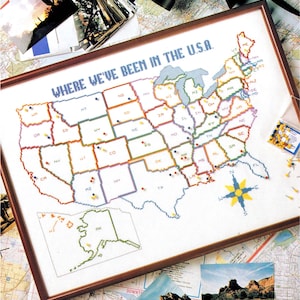 May include: A framed cross-stitch map of the United States with the text WHERE WE'VE BEEN IN THE U.S.A. The map features state abbreviations, colorful borders, a compass rose, and insets for Alaska and Hawaii. Small push pins are inserted into various states to mark locations.