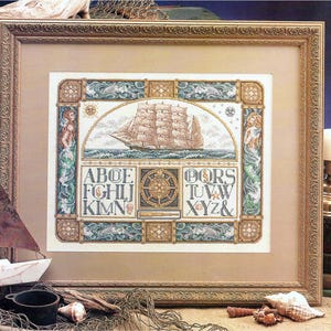 May include: Framed nautical cross-stitch art featuring a tall ship sailing on the sea. The design includes an alphabet, compass rose, and mermaids, all in shades of blue, beige, and gold. The frame is ornate and gold-toned.
