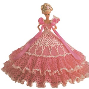 May include: A doll wearing a pink and white crocheted gown. The dress features a layered skirt, a bodice with a decorative openwork design, and pink ribbon accents. The doll has blonde hair styled in an updo with small pink flowers.