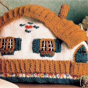 Puede incluir: Un calentador de té tejido con forma de casa. El calentador presenta un cuerpo blanco con techo y ribetes marrones, contraventanas verdes y un diseño de corazón rojo y hojas verdes. El artículo está hecho de hilo y tiene una apariencia texturizada.