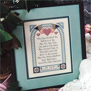 Könnte beinhalten: Gerahmte Kreuzsticharbeit mit einem irischen Hochzeitssegen. Der Text, in blauem Faden, lautet "May God Be With You" und andere Segenswünsche. Das Design umfasst Herzen und dekorative Bordüren, gerahmt in Schwarz.