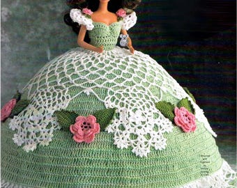 May include: A doll in a handmade green and white crochet dress, adorned with pink flower embellishments. The dress has a detailed lace design and a scalloped edge, reflecting a vintage craft aesthetic.