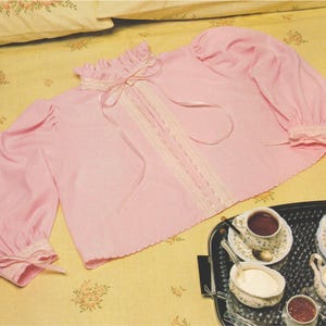 May include: A light pink long-sleeve blouse featuring a ruffled high collar, a vertical lace panel down the center, and matching lace trim at the cuffs. The garment includes thin satin ribbon ties at the neckline and at each of the sleeve cuffs.
