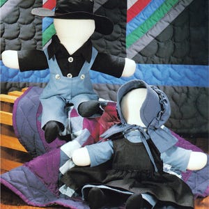 May include: Two fabric dolls with blank faces. One doll wears a black hat, black shirt, and blue overalls. The other doll wears a blue bonnet, a blue long-sleeved shirt, and a black dress. Both dolls have white fabric heads and hands.
