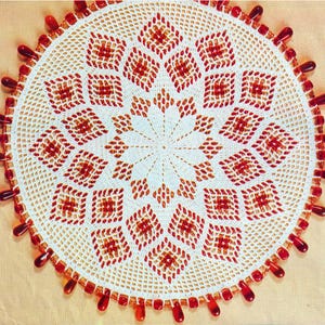 May include: A handmade, white crocheted doily with a central starburst design and repeating diamond patterns in shades of red and brown. The edge is adorned with red teardrop-shaped beads, creating a decorative and intricate piece.