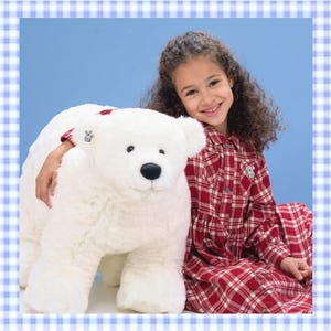 May include: A large, white plush polar bear toy with a black nose and a small metallic button tag on its ear. The bear has soft, textured fur and is positioned next to a person wearing a long-sleeved red and white plaid button-down dress.