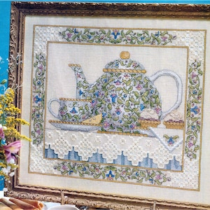 May include: Cross-stitch artwork featuring a teapot and teacup with floral patterns in blue, green, and pink. The framed piece includes a decorative border with similar floral motifs. The artwork is set against a cream-colored background.