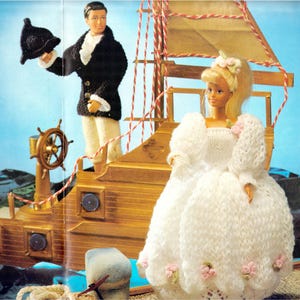 May include: A doll in a white knitted wedding dress with pink floral details stands near a sailor doll on a wooden toy ship. The sailor doll wears a black jacket, white trousers, and holds a black hat. The ship features a gold steering wheel.