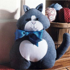 May include: A plush cat figurine made of dark grey fabric with a white heart-shaped patch on its chest and a white face patch. It features black button eyes, a stitched nose and mouth, and a large blue ribbon tied around its neck.