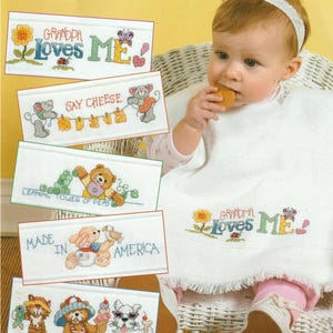 May include: A baby wearing a white bib with the text "GRANDMA Loves ME" embroidered in colorful thread. The image also shows several cross-stitch designs with text and cartoon characters, including "GRANDPA Loves ME" and "MADE IN AMERICA."
