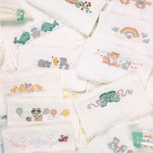 Puede incluir: Un conjunto de toallitas para bebé blancas con diseños de punto de cruz coloridos. Los diseños incluyen animales, arcoíris, globos y las palabras "happy birthday" y "baby".