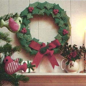 Peut inclure: Une couronne en tissu composée de feuilles vertes à motifs et de petites grappes de baies rouges, ornée d'un grand nœud rouge à la base. Deux décorations d'oiseaux rembourrés sont suspendues, l'un vert clair aux ailes roses et l'autre rose rayé de blanc.