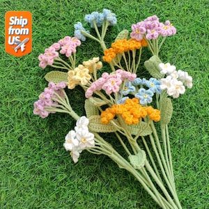 May include: A collection of handmade crochet flowers in shades of pink, yellow, orange, blue, and white. The flowers are arranged with green stems and leaves, set against a backdrop of green grass.