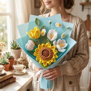 May include: A handcrafted crochet bouquet with a large yellow sunflower, white calla lilies, and daisies, wrapped in light blue paper and tied with a white ribbon. The bouquet includes green leaves and is held by a person.