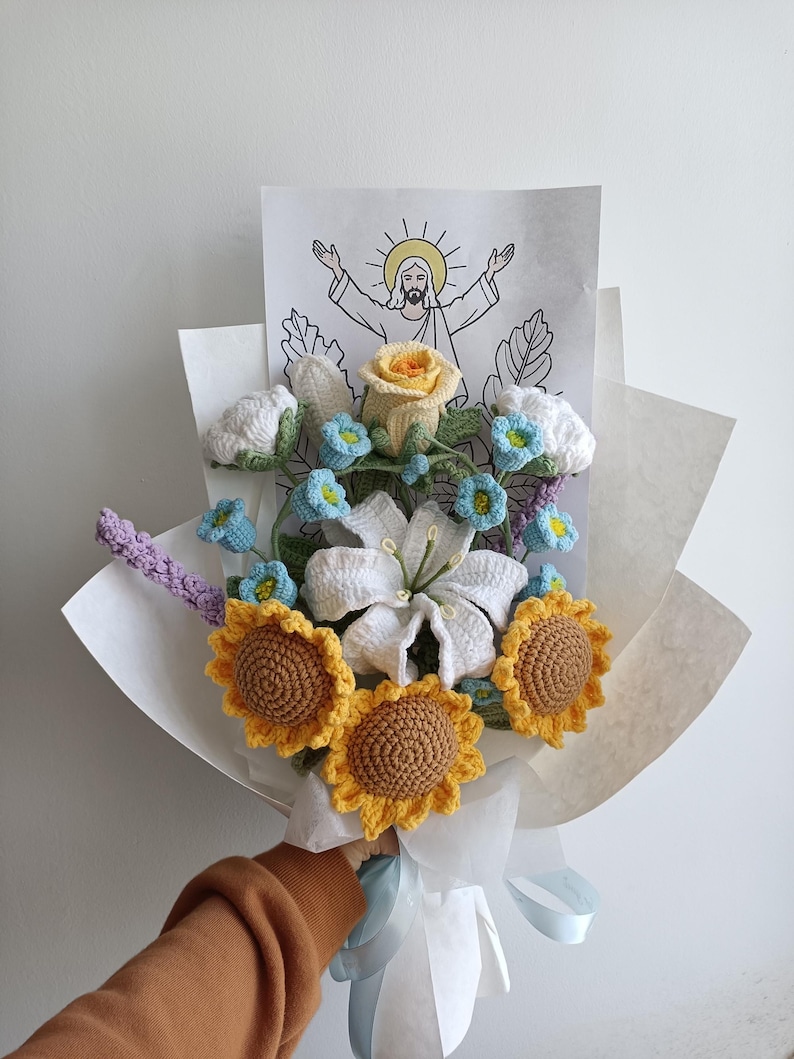 Puede incluir: Un ramo de flores de ganchillo envuelto en papel blanco. El ramo incluye girasoles, lirios, rosas y nomeolvides. Un dibujo de Jes&uacute;s con las palabras "Ha resucitado" es visible en el fondo.
