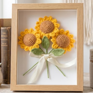 以下が含まれることがあります： 3つの手編みのヒマワリの花束が額装されたアート作品。ヒマワリは黄色い花びらと茶色の中心部を持ち、緑の葉と茎が付いています。クリーム色のリボンで花束が結ばれています。フレームは明るい色の木材で作られています。