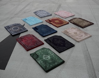 May include: An assortment of prayer mats in various colors, including blue, red, green, and beige, are arranged on a concrete surface. Each mat features an ornate, patterned design. The word "UIT" is visible in the background.