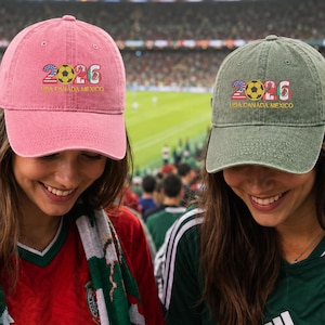 May include: A pink baseball cap and an olive green baseball cap, both featuring the embroidered text 2026 and USA CANADA MEXICO with a soccer ball graphic. Each cap has a curved brim and a structured crown, displaying a casual, sporty design.
