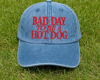 May include: A blue baseball cap with the embroidered phrase "BAD DAY TO BE A HOT DOG" in red. The hat is made of a soft material and has a curved brim. The cap is sitting on a bed of green grass.