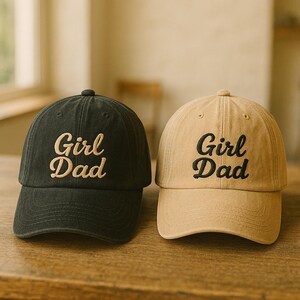 May include: Two baseball caps, one black and one tan, sit side-by-side. Each cap has the embroidered text "Girl Dad" in a contrasting color. The caps are made of a soft fabric and have a curved brim.
