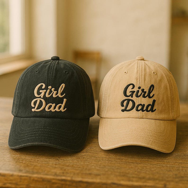 May include: Two baseball caps, one black and one tan, sit side-by-side. Each cap has the embroidered text "Girl Dad" in a contrasting color. The caps are made of a soft fabric and have a curved brim.