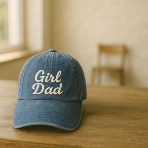 May include: A blue denim baseball cap with the words "Girl Dad" embroidered in white script across the front. The cap has a curved brim and a classic six-panel design. The hat is sitting on a wooden surface.