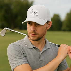 Könnte beinhalten: Eine weiße Baseballkappe mit den Buchstaben "UXM" und gekreuzten Golfschlägern in Schwarz. Die Kappe hat einen gebogenen Schirm. Die Person trägt ein graues Poloshirt.