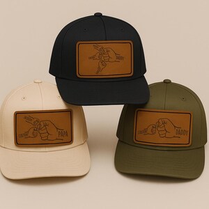 Op de afbeelding: Drie baseballpetten in zwart, crème en olijfgroen. Elke pet heeft een bruin leren patch met een vuist-bump ontwerp en tekst. De zwarte pet zegt "Daddy", de crème pet zegt "Papa" en de olijfgroene pet zegt "Daddy".