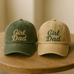 May include: Two baseball caps, one olive green and one tan, are displayed. Both caps feature the embroidered text "Girl Dad" in a contrasting color. The caps have a classic six-panel design with a curved brim and an adjustable strap.