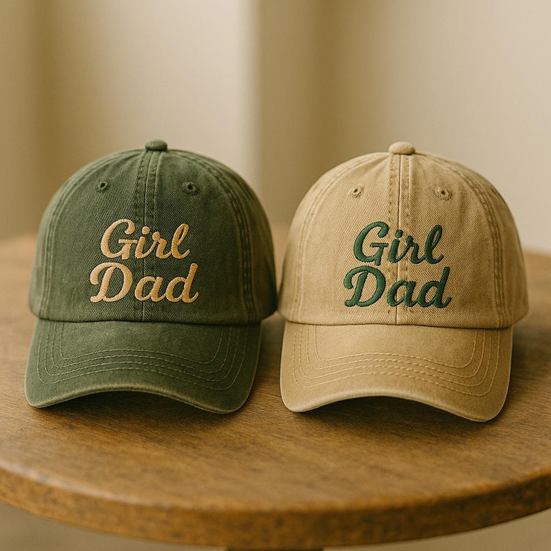 May include: Two baseball caps, one olive green and one tan, are displayed. Both caps feature the embroidered text "Girl Dad" in a contrasting color. The caps have a classic six-panel design with a curved brim and an adjustable strap.