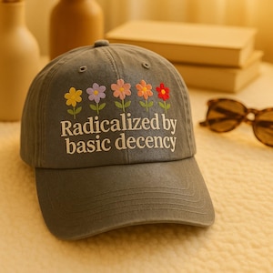 Op de afbeelding: Een grijze baseballcap met geborduurde bloemen in geel, paars, roze, oranje en rood. De tekst "Radicalized by basic decency" is eronder gedrukt. De cap heeft een gebogen klep.