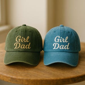May include: Two baseball caps, one olive green and one blue, sit side-by-side on a wooden surface. Each cap has the embroidered text "Girl Dad" in a contrasting color. The caps have a classic, curved brim and a button on top.