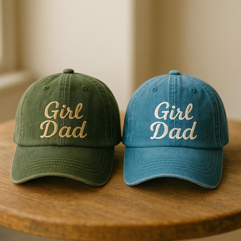 May include: Two baseball caps, one olive green and one blue, sit side-by-side on a wooden surface. Each cap has the embroidered text "Girl Dad" in a contrasting color. The caps have a classic, curved brim and a button on top.