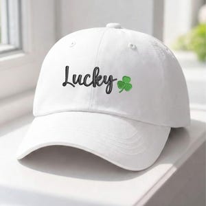 May include: A white baseball cap featuring the word "Lucky" in black script, with a green shamrock emblem. The cap has a curved peak and a classic six-panel design, suitable for everyday wear.