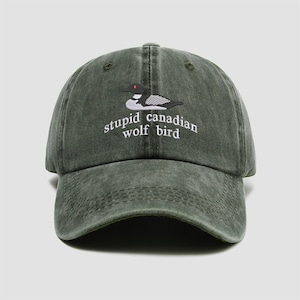 May include: A green baseball cap with a curved brim. The cap features an embroidered design of a bird and the words "stupid canadian wolf bird" in white. The cap is made of a worn, faded material.