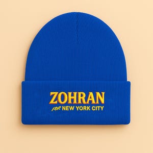 Può includere: Un berretto blu reale con la scritta "ZOHRAN" ricamata in giallo e arancione, con "FOR NEW YORK CITY" sotto. Il cappello è realizzato in un materiale a maglia a coste.
