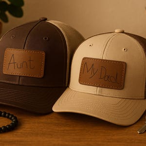 Op de afbeelding: Twee baseballpetten met leren patches. Eén is bruin met "Aunt" op de patch, de andere is beige met "My Dad" op de patch. Een zwarte kralenarmband en sleutels liggen op het houten oppervlak.