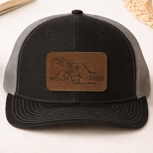 May include: Black and gray trucker hat with a brown leather patch. The patch features a graphic of three fists bumping, with the names "Jackson" and "Kinsley" above and "Grandpa" below. The hat has a curved brim.