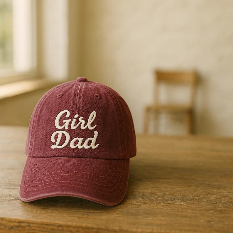 May include: A maroon baseball cap with the embroidered text "Girl Dad" in white script. The cap is made of a soft fabric and has a curved brim. The hat is sitting on a wooden surface.
