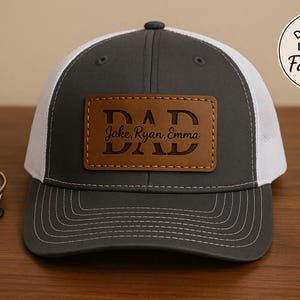 Op de afbeelding: Een grijze en witte truckerpet met een bruin leren patch met de tekst "DAD" met de namen "Jake, Ryan, Emma" eronder. De pet ligt op een houten oppervlak met sleutels en een kom op de achtergrond. Het is een Vaderdagcadeau.