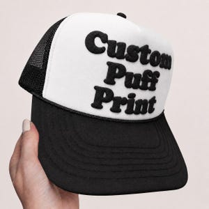 Könnte beinhalten: Eine schwarz-weiße Trucker-Cap mit weißer Schaumstoff-Frontpartie und dem schwarzen Schriftzug Custom Puff Print in erhabener, dreidimensionaler Optik. Die Kappe verfügt über ein schwarzes Netzgewebe, einen schwarzen Schirm mit Nähten und eine schwarze Kordel.