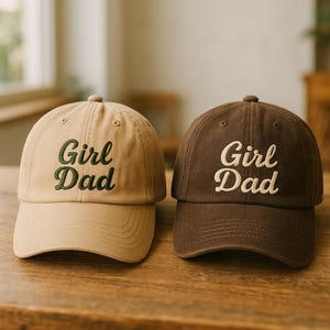 May include: Two baseball caps, one tan and one brown, sit side-by-side. The tan cap has green embroidered text that reads "Girl Dad." The brown cap has white embroidered text that reads "Girl Dad."