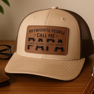 Op de afbeelding: Beige en bruine baseballcap met een leren patch met de tekst "MY FAVORITE PEOPLE CALL ME PAPA" met namen eronder. Een horloge en een bril liggen naast de pet.