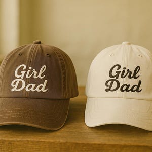 May include: Two baseball caps, one brown and one off-white, sit side-by-side on a wooden surface. Each cap has a curved brim and the words "Girl Dad" embroidered in a cursive font across the front. The caps are casual and stylish.