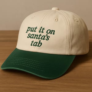 May include: A baseball cap featuring a cream-coloured crown and a dark green brim. The words "put it on santa's tab" are embroidered in green on the front. The cap has a curved peak and a button on top, suitable for casual wear.