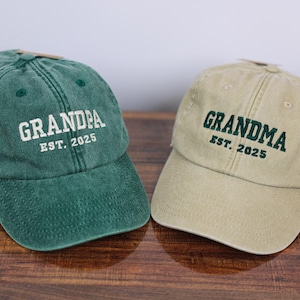 Może przedstawiać: Dwie czapki bejsbolowe, jedna zielona, a druga beżowa, z haftowanym tekstem. Zielona czapka ma napis "Grandpa Est. 2025", a beżowa czapka ma napis "Grandma Est. 2025".