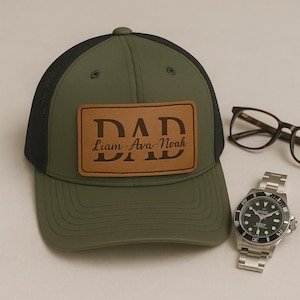 Może przedstawiać: Zielono-czarna czapka bejsbolówka z brązowym skórzanym naszywką z napisem "DAD" i nazwiskami "Liam, Ava, Noah" pod spodem.